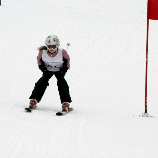 Ein junger Skifahrer, der einen Helm und eine Brille trägt, navigiert eine verschneite Piste. Die Kleidung des Skifahrers besteht aus einem weißen und rosafarbenen Weste mit Logo. Sie fahren an einem roten Flaggen vorbei.