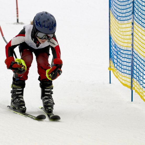 Ein Skifahrer mit blauem Helm und roter Skiausrüstung lehnt sich auf schneebedeckten Hängen nach vorne, hält Skistöcke in den Händen und hat ein blaues und gelbes Netz im Hintergrund.
