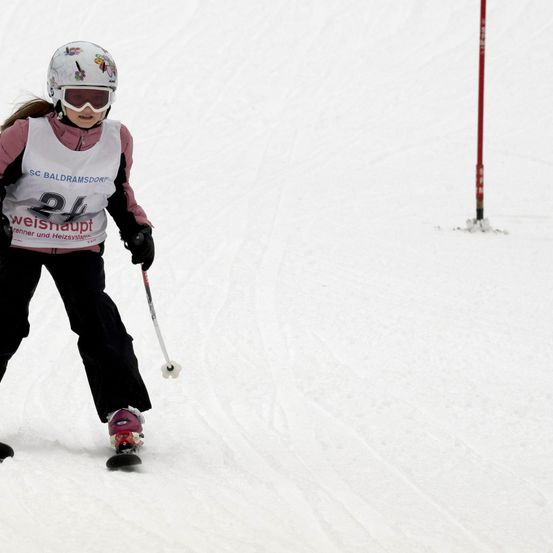 Eine junge Skifahrerin fährt einen schneidigen Hang hinunter. Sie trägt einen Helm, eine Brille und eine Startnummer 24. Sie hält einen Skistock in der rechten Hand.