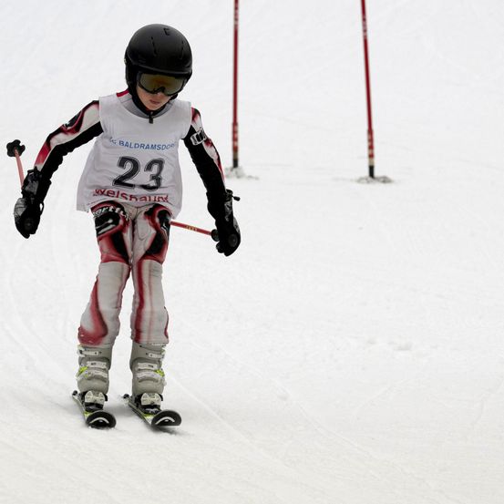 Ein junger Skifahrer mit der Nummer 23 fährt mit Schutzausrüstung einen verschneiten Hang hinunter.