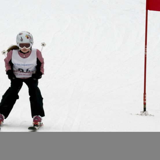 Ein Skifahrer mit weißem Helm und Schutzbrille fährt einen schneebedeckten Hang hinunter. Ein roter Flaggenpfosten steht rechts im Schnee.