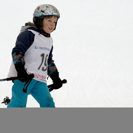 Ein junger Skifahrer mit der Nummer 79 auf seinem weißen Startnummern-Trikot fährt bergab. Er trägt einen Helm, Handschuhe und Skistöcke.