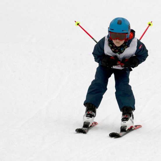 Ein junger Skifahrer mit blauem Helm und Skibrille fährt auf einer verschneiten Piste. Er hält Skistöcke in beiden Händen.