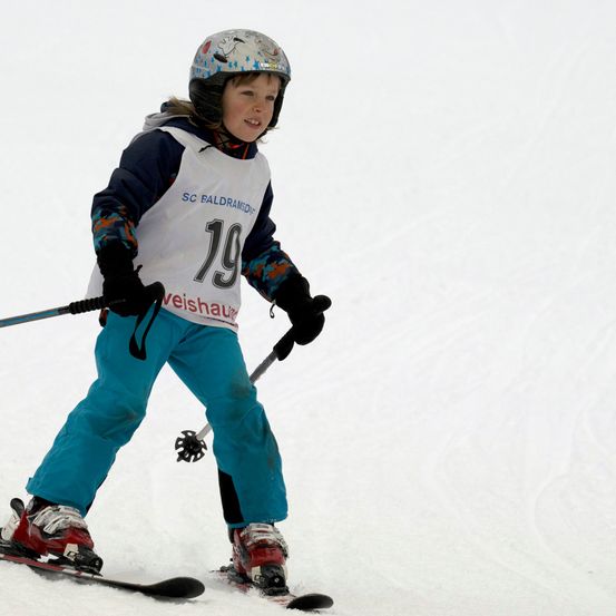Ein junger Skifahrer mit Helm und Skianzug mit der Nummer 19 fährt einen schneebedeckten Hang hinunter.