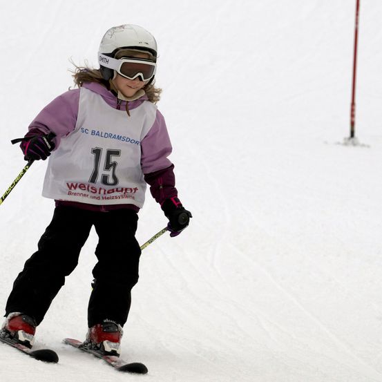Ein junger Skifahrer, der einen weißen Helm und eine Schutzbrille trägt, hält Skistöcke in der Hand und fährt einen schneebedeckten Hang hinunter. Er trägt eine weiße und lila Jacke mit der Nummer 15.