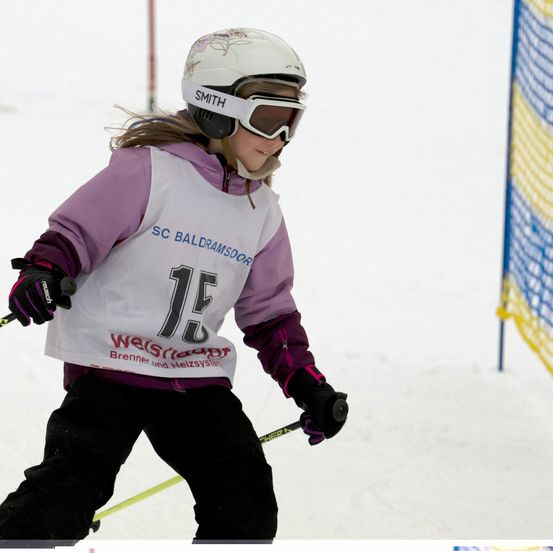 Ein junges Mädchen, das den Berg hinunterskit und einen weißen Helm, eine Brille und eine nummerierte Startnummer mit SC BALDRAMSDORR trägt. Die Nummer ist 15.