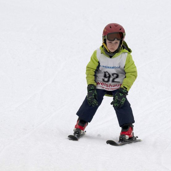 Ein Kind auf Skiern mit rotem Helm, Schutzbrille und nummerierter Skijacke fährt auf einem verschneiten Hang.