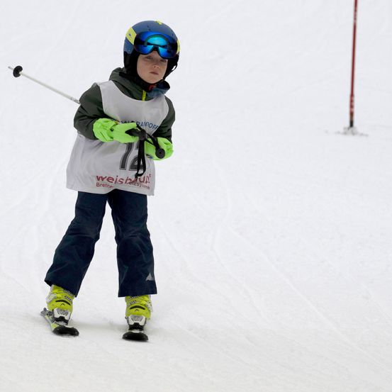 Ein junger Skifahrer in einem blauen Helm und Schutzbrille fährt einen schneebedeckten Hang hinunter, trägt eine weiße Weste mit der Nummer 14 und grüne Handschuhe.