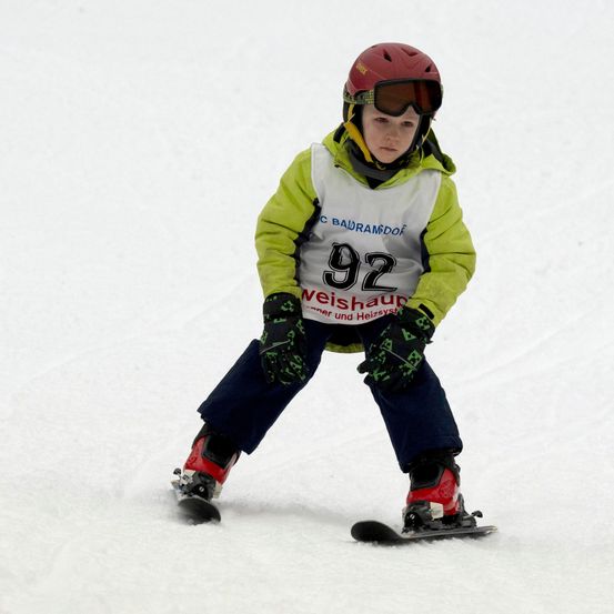 Ein junger Junge snowboardet einen schneebedeckten Hang hinunter, trägt einen roten Helm, eine Schutzbrille und eine grün-weiße Jacke. Er hat eine Nummer 92 auf der Brust und hält Skistöcke.