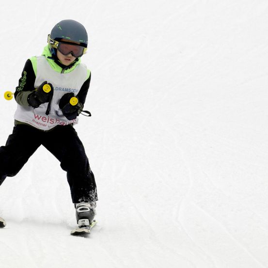 Ein junger Skifahrer mit schwarzem Helm und Schutzbrille fährt einen schneebedeckten Hang hinunter. Der Skifahrer trägt eine grüne Weste mit weißem Text und der Nummer 3.