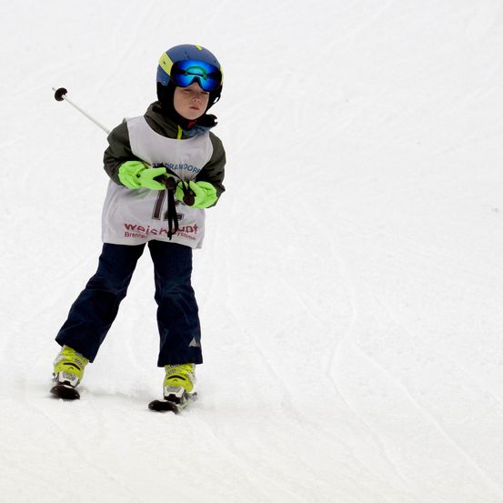 Ein junger Skifahrer, der eine Schutzbrille und einen Helm trägt, fährt den Schneehang hinunter. Sie halten einen Skistock in der rechten Hand.