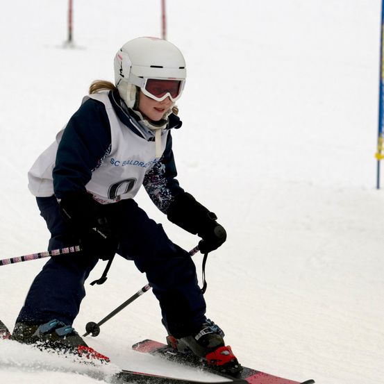 Ein junges Mädchen fährt auf einem verschneiten Hang Ski. Sie trägt einen weißen Helm, eine Brille, eine Jacke mit 'SC BALDRIDGE' auf der Vorderseite und blaue Skihosen.