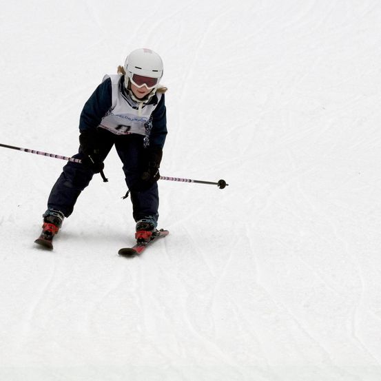 Ein junger Skifahrer in blauer Skiausrüstung fährt mit Skistöcken in der Hand einen verschneiten Hang hinunter.