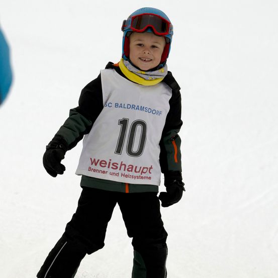 Ein Kind, das ein weißes Hemd mit der Nummer 10 und SC Baldramsdorf darauf trägt, steht auf Schnee und trägt eine Schneebrille und einen Helm.