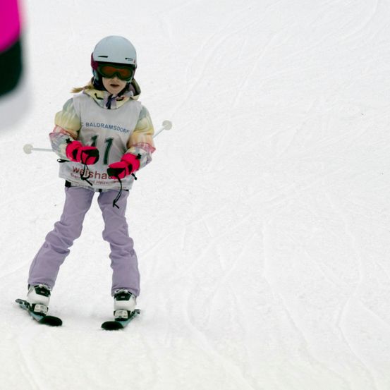 Ein junges Mädchen in Skiausrüstung, Helm und Schutzbrille, fährt den Schneehang hinunter. Sie trägt ein farbenfrohes Skioutfit mit der Nummer 11 und hält Skistöcke.