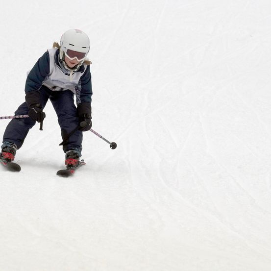 Ein junges Kind, das einen schneebedeckten Hang hinunterskit, trägt einen weißen Helm, eine Brille und blaue Skiausrüstung und hält Skistöcke in der Hand.