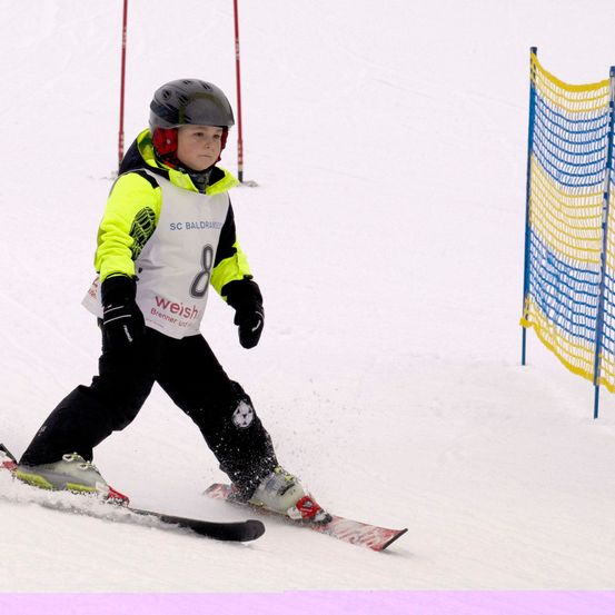 Ein junger Skifahrer, Nummer 8, in Helm und Skiausrüstung, fährt einen schneebedeckten Hang hinunter. Schnee spritzt um ihn herum.