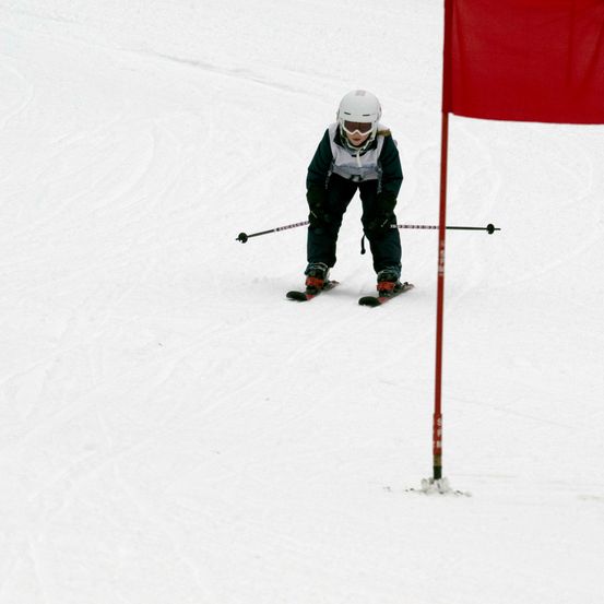 Ein Skifahrer bewegt sich auf einer schneebedeckten Piste und passiert eine rote Fahne an einem Pfosten. Der Skifahrer trägt einen weißen Helm, eine Brille und eine dunkle Jacke.
