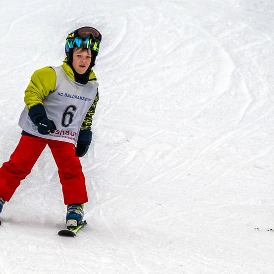 Ein junger Junge, der auf einem verschneiten Hang Ski fährt, trägt eine gelbe und grüne Jacke mit der Nummer 6, einen Helm und rote Hosen. Er hält einen Skistock in der rechten Hand.