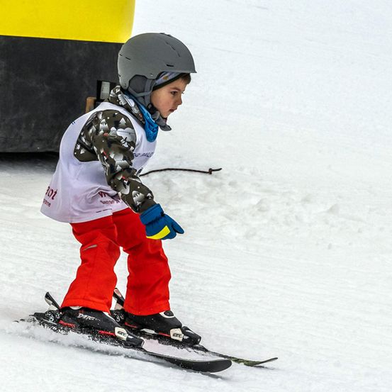 Ein junger Skifahrer mit Helm, Handschuhen und roten Skihosen fährt einen verschneiten Hang hinunter. Hinter ihm ist ein gelber und schwarzer Schneepflug zu sehen.