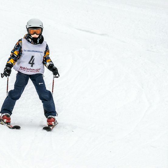 Ein Skifahrer in einem weißen Helm und mit Skibrille fährt einen verschneiten Hang hinunter, trägt ein nummeriertes Trikot und hält Skistöcke.