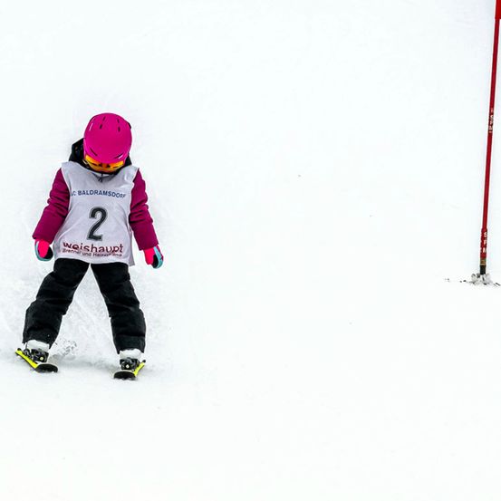 Ein Kind in einem Skianzug mit der Nummer 2 darauf, das einen verschneiten Hang hinunterfährt, mit einem roten Pfosten rechts.