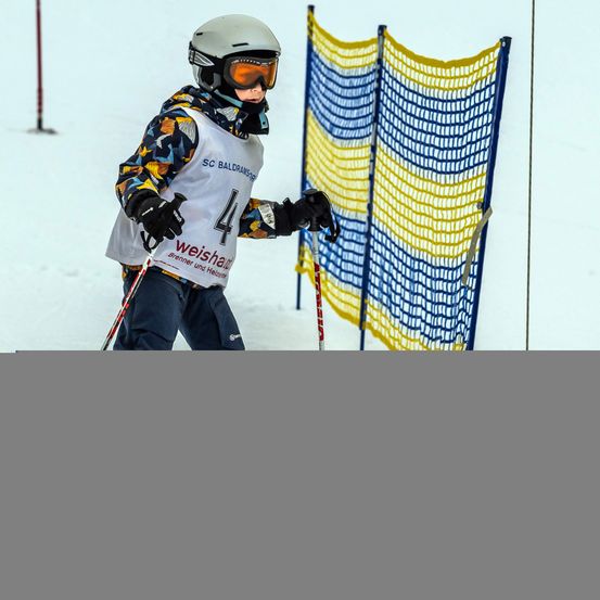 Ein Skifahrer in einem weißen Trikot mit der Nummer 44 steht auf einem verschneiten Hang, trägt eine Schutzbrille und hält Skistöcke. Ein blaues und gelbes Netzhindernis steht hinter ihm.