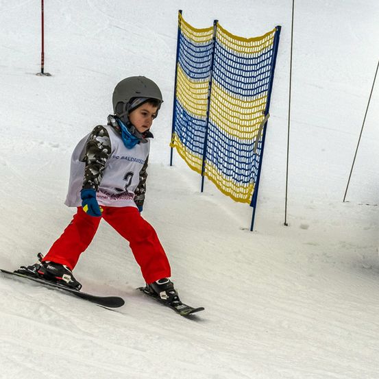 Ein junger Junge in einem Helm und Skiausrüstung fährt einen verschneiten Hang hinunter, trägt eine weiße und graue Jacke mit einem blauen Schal.