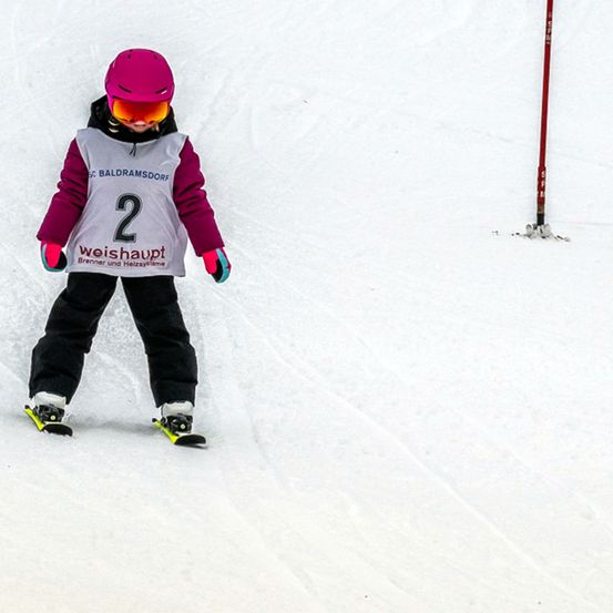 Ein Kind beim Skifahren auf einer verschneiten Piste, das ein weißes Hemd mit der Nummer 2 und eine lila Jacke trägt.