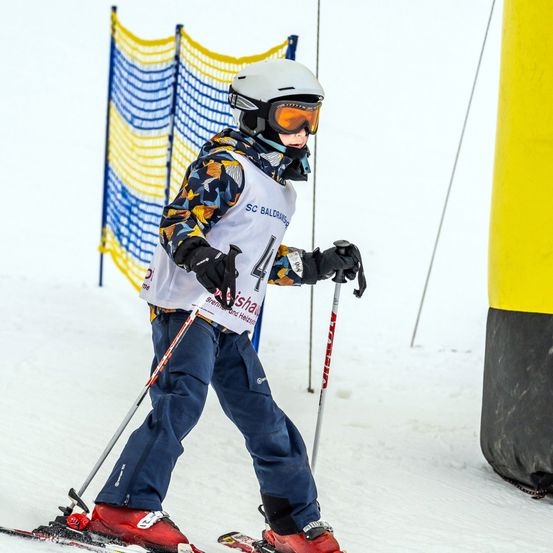 Ein junger Skifahrer in einem weißen Helm, mit Brille und Skianzug mit der Nummer 44 darauf, fährt einen verschneiten Hang hinunter und hält Skistöcke. Dahinter befindet sich ein blau-gelbes Netzgitter.