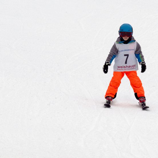 Ein Skifahrer mit blauem Helm und der Nummer 7 steht auf einem verschneiten Hang, trägt orangefarbene Hosen und Handschuhe.