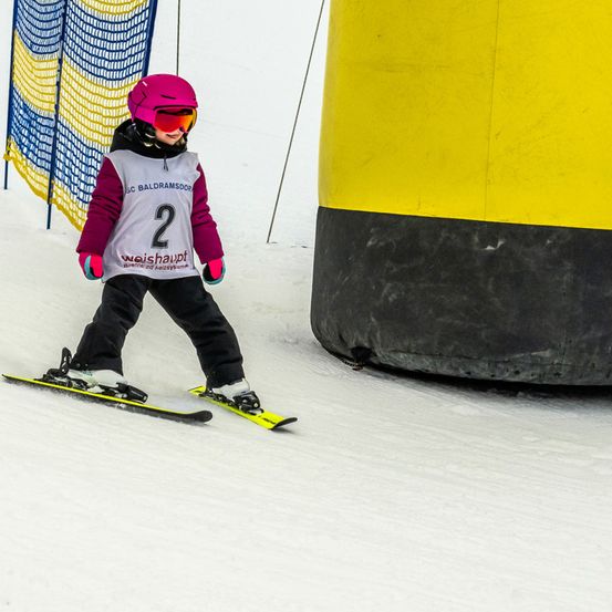 Ein Kind mit pinkem Helm fährt mit Skiern bergab und passiert ein gelbes und schwarzes Hindernis. Es trägt eine Nummernweste und eine Brille.