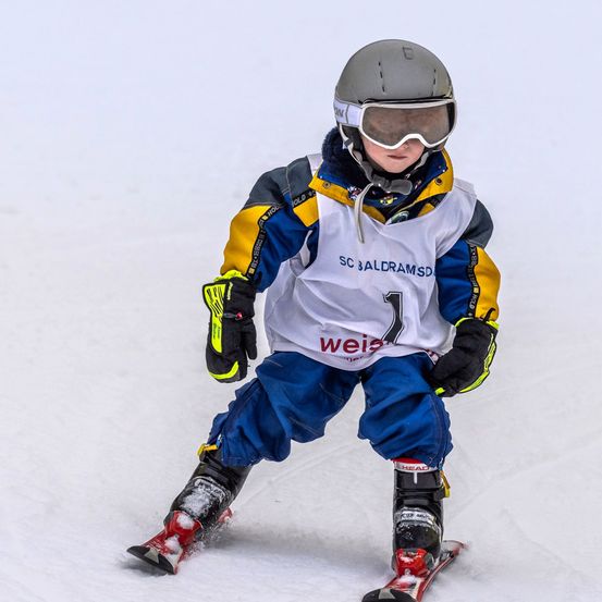 Ein junger Skifahrer in voller Ausrüstung, einschließlich Helm, Brille und Handschuhen, fährt einen schneebedeckten Hang hinunter. Der Skifahrer trägt eine weiß-gelb-blaue Jacke mit der Nummer 1 und dem Wort 'weis' in prominenter Platzierung.