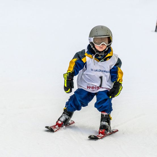 Ein junger Skifahrer in einem weißen und blauen Jackett mit SC Baldramsd darauf, der einen verschneiten Hang hinunterfährt.