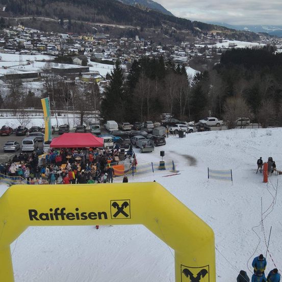 Eine verschneite Landschaft mit einer Menschenmenge, die sich um ein rotes Zelt versammelt. Im Vordergrund befindet sich ein gelber Bogen mit dem Wort Raiffeisen.