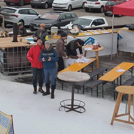 Eine verschneite Außenfläche mit mehreren geparkten Autos. Ein Mann in einem roten Mantel und eine Frau in einem blauen Mantel stehen vor einem Tisch. Ein anderer Mann beugt sich über einen Tisch mit Gegenständen. Dahinter stehen Holzbänke, ein Tisch mit einem roten Zelt und andere Gegenstände.