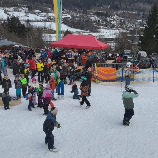 Ein verschneiter Bereich mit einem roten Zelt und vielen Menschen, einige mit Mützen und Handschuhen, andere beim Skifahren und andere zuschauend. Ein Auto ist in der Ferne geparkt.