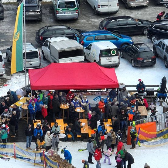 Eine Draufsicht auf eine Menschenmenge, die sich um ein rotes Zelt und Tische in einem verschneiten Bereich versammelt. Ein Flaggenmast steht neben dem Zelt. Einige Menschen tragen Mützen und Stiefel.
