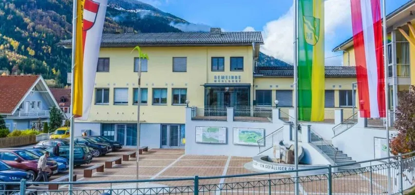 Ein gelbes Gebäude namens Gemeindehaus mit Fahnen davor. Mehrere Autos sind vor dem Gebäude geparkt. Bäume und Berge sind im Hintergrund.