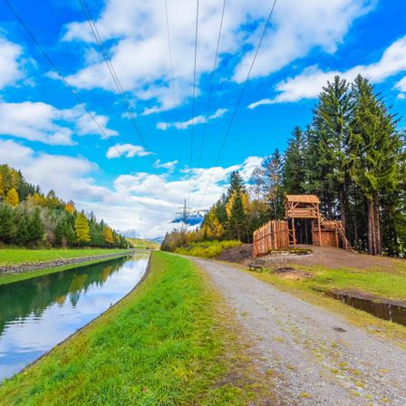 Ein malerischer Weg neben einem Kanal unter einem blauen Himmel mit Wolken. Es gibt eine Holzstruktur in der Nähe des Kanals und einen Holzzaun an der Seite.