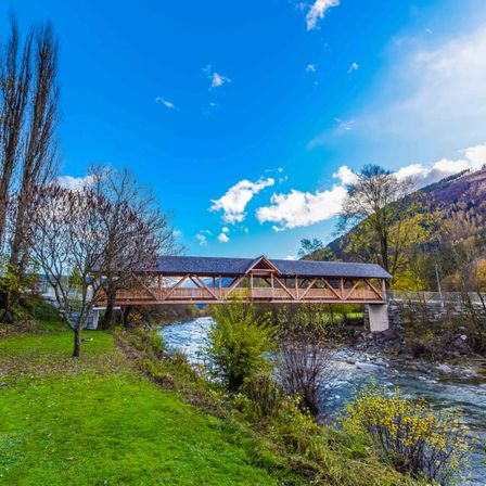 Eine Holzbrücke überspannt einen Fluss mit üppigem Grün und Bergen im Hintergrund unter einem blauen Himmel mit Wolken.
