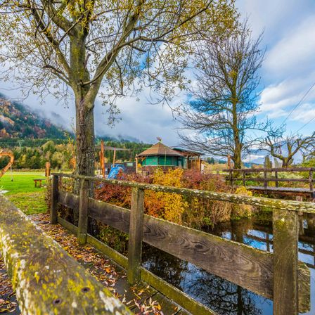 Ein Holzzaun umgibt einen kleinen Teich mit einem Pavillon im Hintergrund. Vorne sind Bäume und Pflanzen zu sehen, und ein grünes Feld ist sichtbar.
