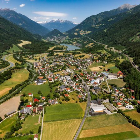 Luftaufnahme eines Dorfes zwischen Bergen, mit einem Fluss und üppiger Grünfläche. Gebäude unterschiedlicher Größe und Stil sind verstreut.