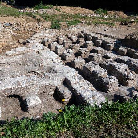 Ein archäologischer Fundort mit großen Steinstrukturen, teilweise vergraben, umgeben von Erde und Gras. Das Gebiet ist uneben, mit einigen Steinen, die aus dem Boden hervorragen.