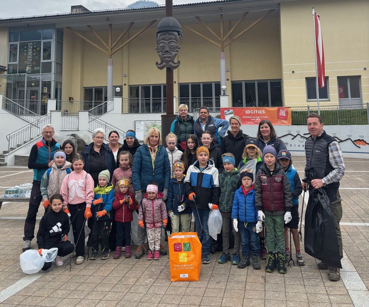 Eine Gruppe von Kindern und Erwachsenen posiert für ein Foto im Freien mit einem Banner im Hintergrund, einer Statue und einem Gebäude. Einige halten Stöcke und Handschuhe.