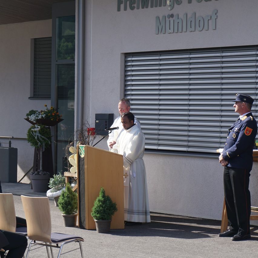 Zwei Männer in formeller Kleidung stehen neben einem Podium. Ein Mann spricht, während ein anderer zuhört. Ein dritter Mann steht in Uniform in der Nähe.