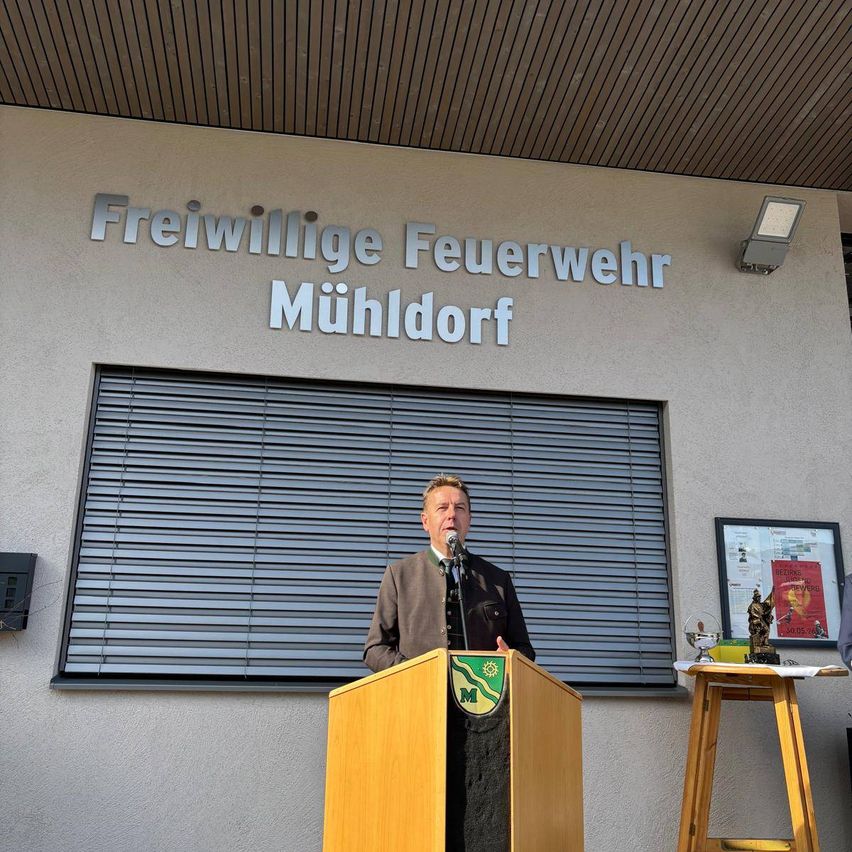 Ein Mann steht vor einem Podium und hält eine Rede vor einem Gebäude mit dem Schild Freiwillige Feuerwehr Mühldorf.
