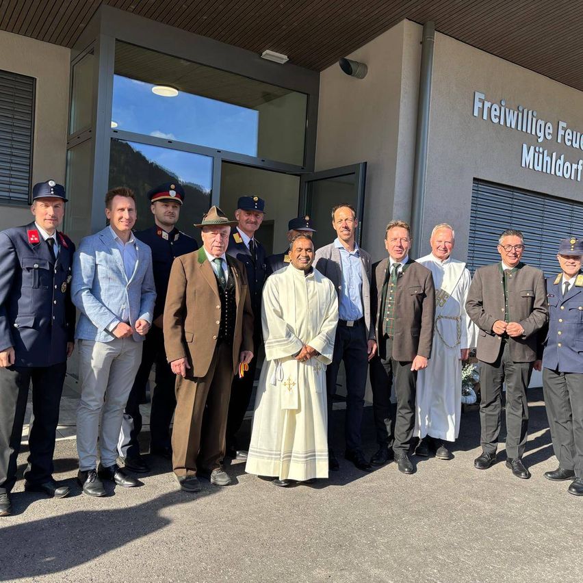 Eine Gruppe von Männern in Uniformen und Anzügen steht vor einem Gebäude mit 'Freiwillige Feuerwehr Mühldorf' an der Wand.