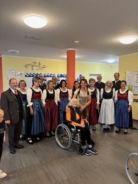 Eine Gruppe von Menschen in traditioneller Kleidung in einem Raum, mit einer Frau im Rollstuhl, lächelt für ein Foto.