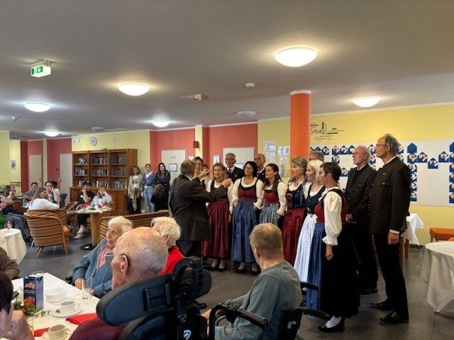 Eine Gruppe von Menschen in traditionellen Kostümen singt ein Lied in einem Raum mit Tischen, Stühlen und einer Bühne.
